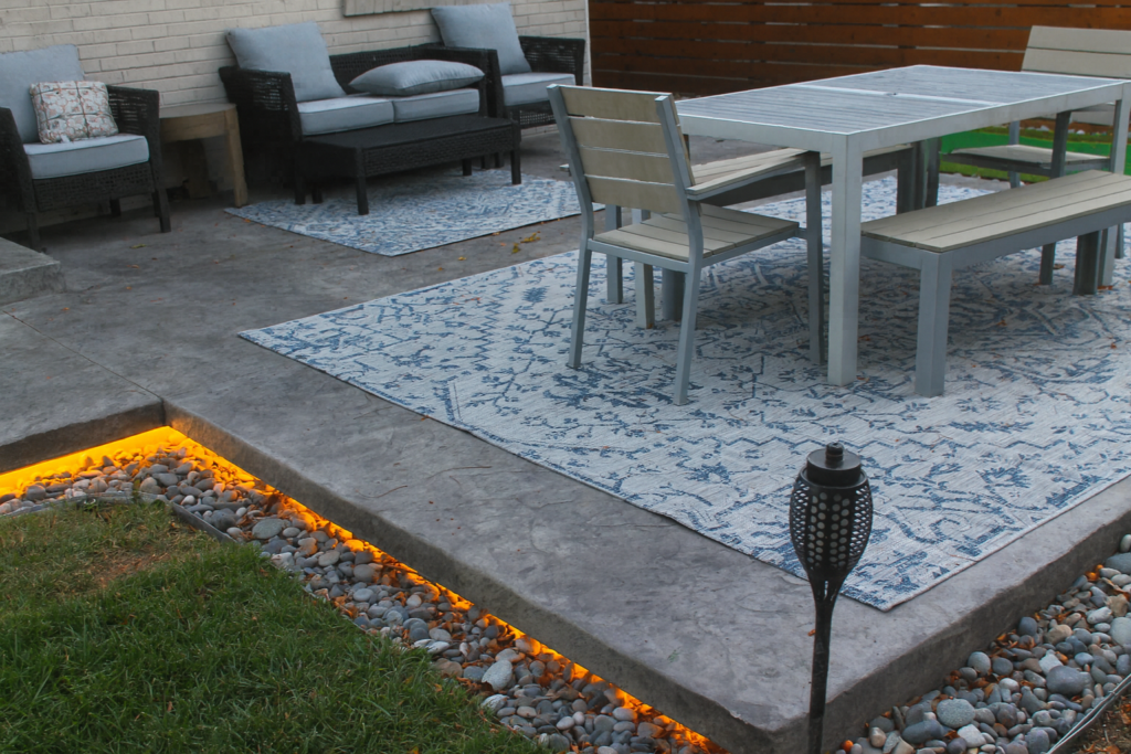 Close view of a stamped concrete patio in a Colorado backyard, showing warm LED edge lighting above a river rock border, outdoor rug and dining set.